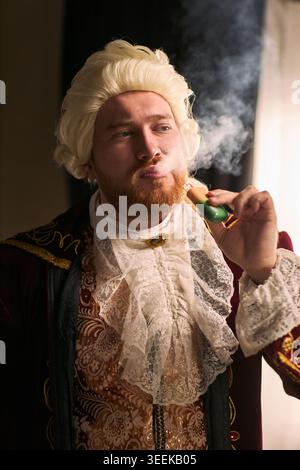 Vertical shot of young man wearing historical costume, exhaling vapor from electronic cigarette and parodying aristocratic lifestyle. Ideal for vaping satire, addiction concept, social commentary Stock Photo