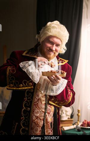 Portrait of young man wearing historical costume, holding fried chicken and smiling at camera. Conceptualizing indulgence, parody, food advertising, vertical Stock Photo