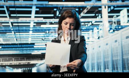 Data center manager verifying infrastructure status with clipboard files and holographic visuals. Server room worker using audit charts and blue wireframe simulation to oversee equipment Stock Photo