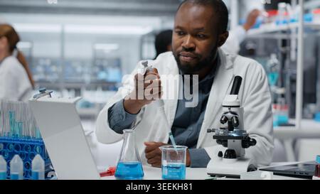 Assay development specialist pipetting reactant into blue solution for purity analysis. Man in research facility lab adding liquid agent to test tube to observe chemical reaction properties, camera B Stock Photo