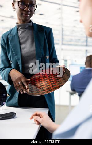 Woman inspecting silicon wafer substrates in industrial plant RD office. Female technician analyzing semiconductor slice in PCB production fab workplace next to coworkers using PCs Stock Photo