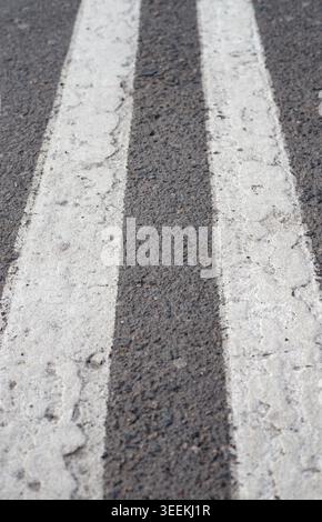Double stripes of white road markings with cracks on dark asphalt. Gray road surface texture and details of a city street with signs of wear and tear. Stock Photo