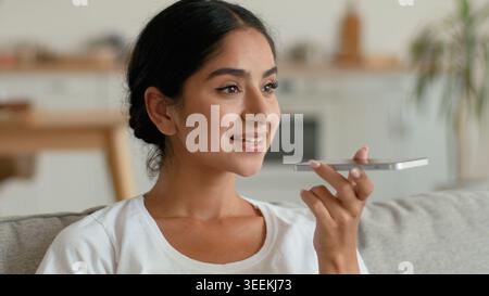 Happy smiling Indian Arabian woman recording voice message mobile phone at home in living room holding smartphone female girl talking into microphone Stock Photo