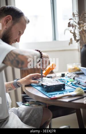 Man works on repairing electronics at home during the day with tools and materials scattered around the table Stock Photo