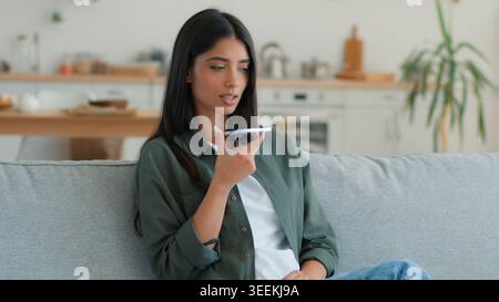 Serious Muslim Arabian woman at home couch recording voice message talking smartphone using loudspeaker mobile phone dictating speech Islamic female Stock Photo