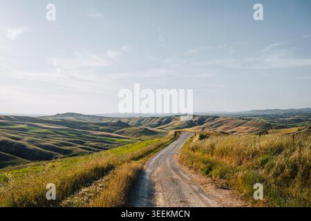 Italy, Tuscany, Crete Senesi: A dirt road winds through rolling hills under a blue sky.. Stock Photo