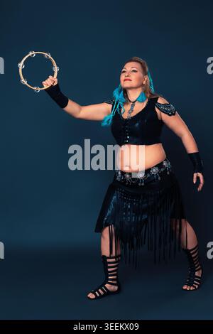 Curvy woman performs with tambourine. Female dancer attired in black ensemble that has been crafted by hand. Outfit consists of leather top and skirt, Stock Photo