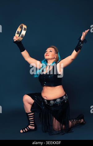 Female dancer stands on one knee with her arms raised up, dances with tambourine and looks up. Mature woman dressed in handmade performance costume - Stock Photo