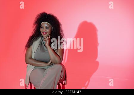Mid-adult African American woman posing on stool in studio in beige outfit, headband, hoops, shadow Stock Photo