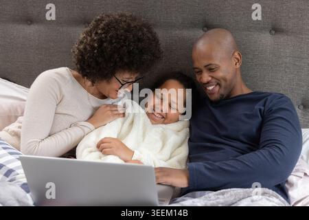 African American family sitting in bedroom on bed, child wearing plush white robe using laptop Stock Photo