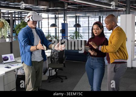 Diverse coworkers guiding senior man wearing white VR headset while referencing tablet at office Stock Photo