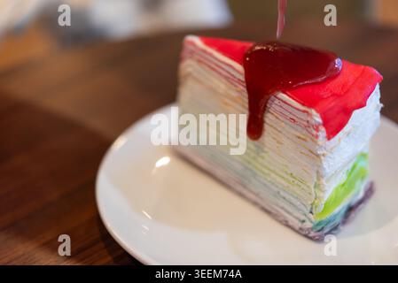 Colorful rainbow crepe cake topped with strawberry sauce and cream served on white plate with soft sweet texture and vibrant dessert presentation Stock Photo