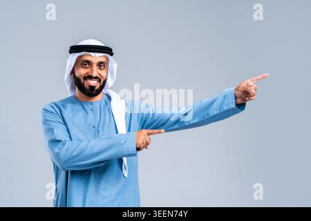 Smiling arab man in traditional kandura and ghutra pointing to the side, indicating a direction or presenting an imaginary product or text, posing in Stock Photo