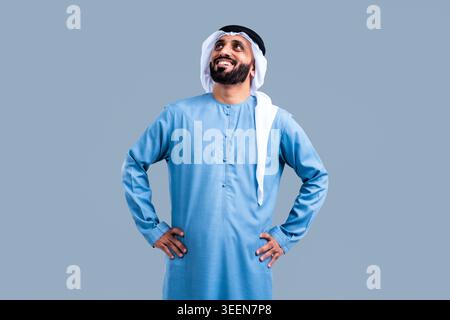 Emirati man in traditional kandura and ghutra, smiling confidently with hands on hips, looking up with optimistic, successful expression against a neu Stock Photo