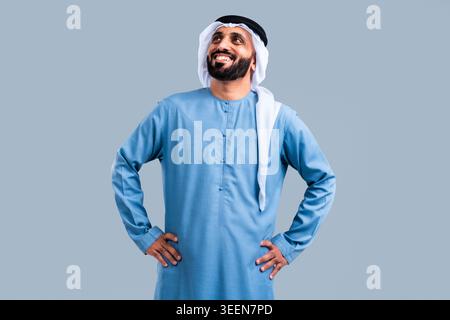 Happy young middle eastern man wearing traditional kandura and ghutra, standing confidently with hands on hips, smiling and looking up, against a simp Stock Photo