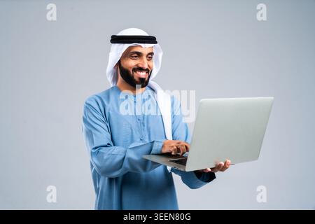 Emirati man wearing traditional clothing, smiling while holding and typing on a laptop, representing global connectivity, modern technology, and succe Stock Photo