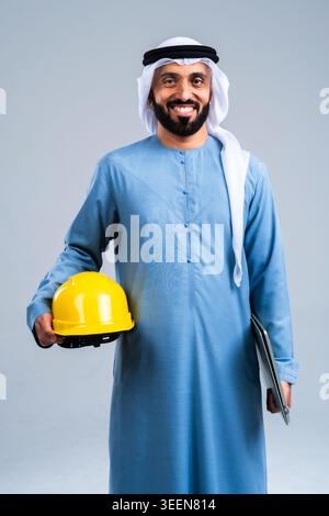 Emirati professional man confidently smiling, holding yellow hard hat and digital tablet, representing professionalism and expertise in engineering, a Stock Photo