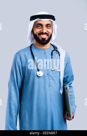 Emirati male doctor smiling, wearing kandura and ghutra, having a stethoscope around his neck, and holding a laptop, representing healthcare, technolo Stock Photo