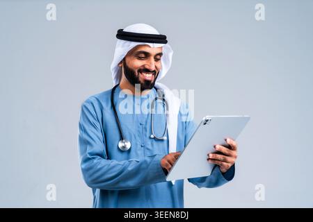 Middle eastern man wearing traditional emirate clothing and a stethoscope, smiling while using a digital tablet, representing modern healthcare, medic Stock Photo