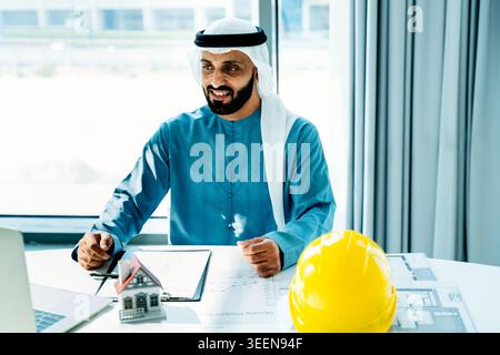 Emirati engineer smiling while working on architectural plans and property models in an office, symbolizing gulf region business, investment, and urba Stock Photo