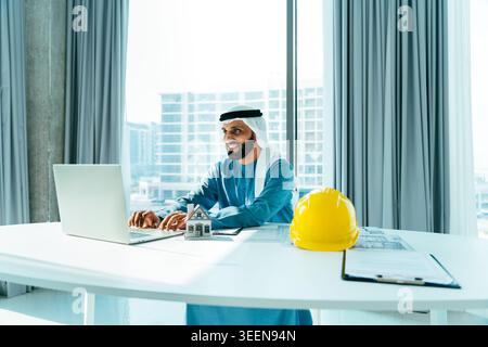 Arab businessman in traditional dress smiling, typing on laptop, reviewing architectural plans with a miniature house and safety helmet on his office Stock Photo