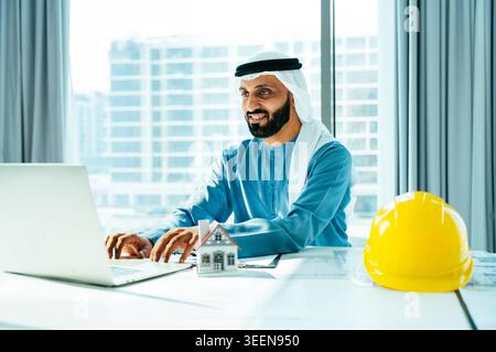 Emirati businessman in traditional kandura smiles while typing on a laptop at a modern office desk with a model house and yellow hard hat, planning a Stock Photo