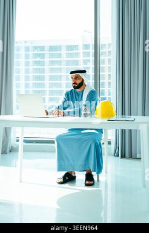 Emirati businessman wearing kandoora and ghutra working on a laptop, analyzing construction projects with a hard hat and house model on a white desk i Stock Photo