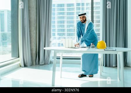 Emirati businessman in kandura and ghutra at office desk, typing on laptop beside hard hat and house model, symbolizing construction, real estate deve Stock Photo