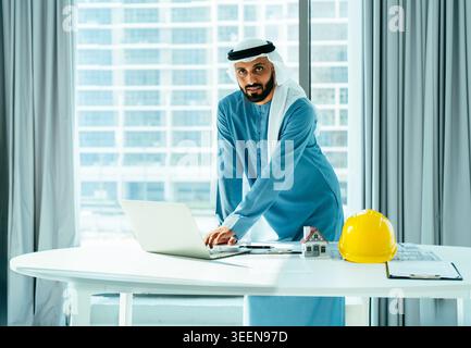 Arab businessman wearing traditional emirati clothing standing at a desk in a modern office, typing on a laptop with a hard hat and house model, focus Stock Photo