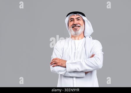 Senior mature emirati man smiling, wearing traditional kandora and ghutra, looking up with arms crossed, portraying confidence, wisdom, and an optimis Stock Photo