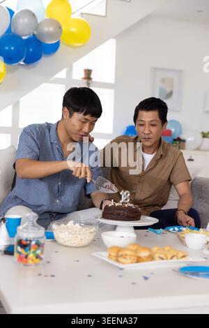 Two Asian men sitting on couch in bright living room cutting chocolate cake using cake server Stock Photo