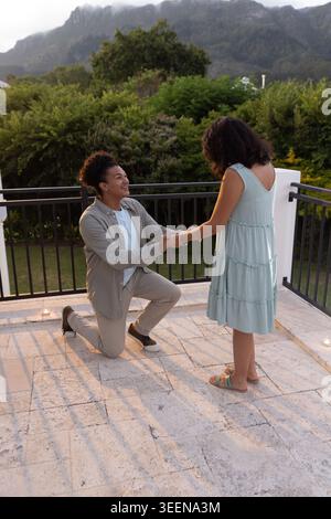Diverse couple kneeling, holding hands on terrace at dusk with candles, woman wearing mint dress Stock Photo