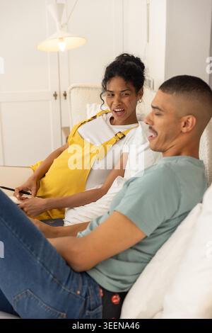 Couple reclining on bed in bedroom, woman in mustard overalls holding slim phone, man laughing Stock Photo
