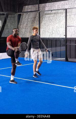 Diverse male teammates stretching and warming up on blue padel court with rackets and mural Stock Photo