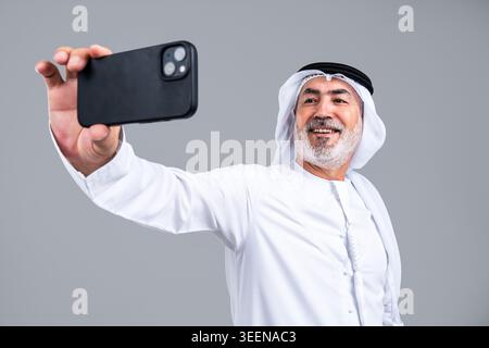 Senior emirati man in kandora and ghutra smiling as he holds a smartphone for a selfie against a muted background, symbolizing connection, technology Stock Photo