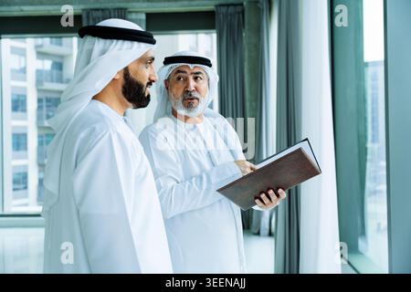 Two middle eastern businessmen wearing traditional emirati clothing are having a serious discussion while one is holding and pointing to documents nea Stock Photo