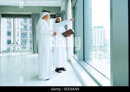 Two emirati businessmen wearing traditional kandoras are standing by a large office window, pointing towards a city view and discussing future investm Stock Photo