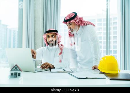 Two saudi businessmen in thobe and ghutra review blueprints and a laptop in a modern office, collaborating on real estate development, planning and in Stock Photo