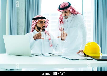 Two saudi men in traditional attire collaborating on architectural blueprints and construction project details, symbolizing corporate teamwork and inv Stock Photo