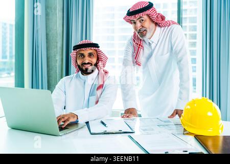 Arab businessmen collaborating on architectural plans and a laptop in a modern office, developing real estate ventures and fostering corporate teamwor Stock Photo