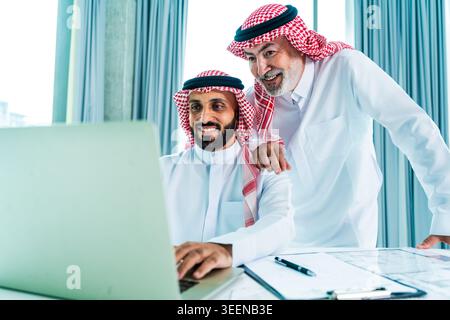 Two saudi business executives in traditional dress collaborate over a laptop in a modern office, planning corporate real estate development with smile Stock Photo