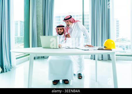 Saudi businessmen in a modern office reviewing blueprints and a laptop, discussing real estate and construction plans, teamwork and strategy for devel Stock Photo