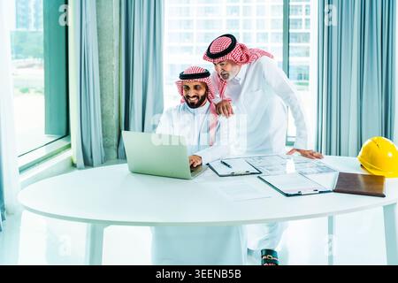 Two saudi businessmen working together on a laptop and reviewing architectural blueprints and documents in a modern office, planning an upcoming const Stock Photo