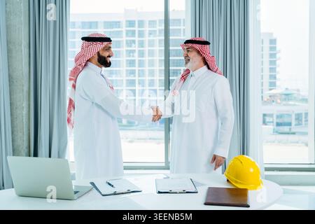 Two saudi businessmen shaking hands in a modern office, symbolizing partnership, agreement, and success in corporate real estate or construction inves Stock Photo
