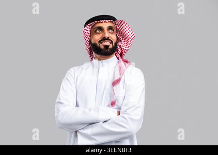 Saudi man wearing traditional ghutra and thobe, smiling confidently while looking upwards, posing with crossed arms in a studio setting on a grey back Stock Photo