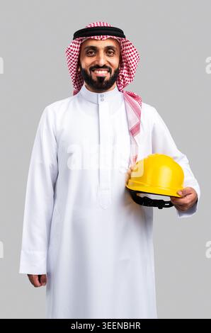 Saudi man standing in traditional thobe and ghutra, holding a yellow hard hat and smiling, representing concepts of professionalism, engineering, and Stock Photo