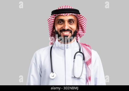 Saudi male doctor standing composed, wearing traditional dishdasha and ghutra, smiling brightly while having a professional stethoscope draped around Stock Photo
