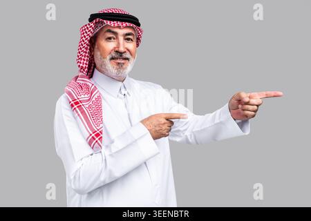 Saudi senior man wearing traditional ghutra and thobe, smiling confidently while pointing fingers to the right, presenting a concept, idea, or informa Stock Photo