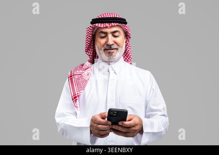 Saudi senior man wearing traditional ghutra and thobe smiling, holding and looking down at a mobile phone, representing connection and modern communic Stock Photo
