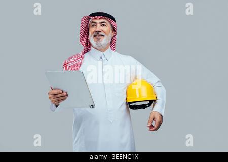 Saudi senior man, dressed in traditional thawb and ghutra, smiling and looking up while holding a digital tablet and yellow hard hat, representing con Stock Photo
